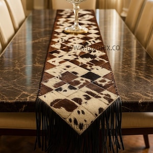 May include: A brown and white cowhide table runner with a diamond pattern and black fringe. The runner is placed on a dark brown marble table. A decorative glass goblet sits on the runner. The website www.fazmax.co is visible.