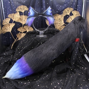 Puede incluir: Una cola peluda negra y azul con un degradado de azul a morado en la punta. La cola se muestra sobre un fondo negro con purpurina dorada. Una diadema peluda negra y azul a juego con orejas se muestra en un soporte en el fondo.