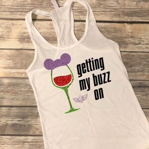 May include: White tank top with a wine glass design. The wine glass has a purple Mickey Mouse ear hat, red wine, and a green stem. The text "getting my buzz on" is printed in black, with a small Buzz Lightyear graphic.