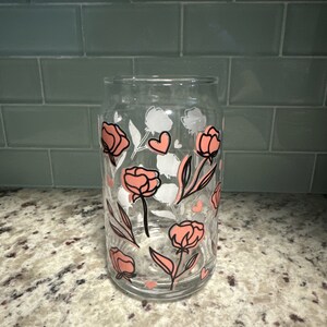 May include: A clear glass drinking glass with a floral pattern. The pattern features pink flowers with black outlines and white flowers with black outlines. There are also small pink hearts in the pattern.