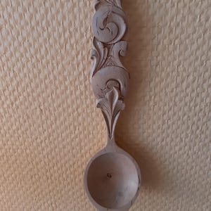 May include: A wooden spoon with a carved, ornate handle. The handle is decorated with intricate floral designs. The spoon is made of light-colored wood and has a smooth, rounded bowl.