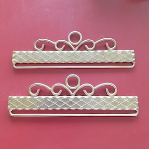 May include: Two gold-colored metal decorative clasps with an ornate, scrolled design. Each clasp features a rectangular center with a diamond pattern and a curved, decorative top. The clasps are displayed against a red background.