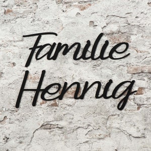 Könnte beinhalten: Schwarzes Metallschild mit den Worten "Familie Hennig" an einer weißen Backsteinwand.