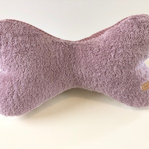 May include: A plush, mauve-colored dog bone shaped pillow. The pillow has a brown tag with the word "Made By" on it.