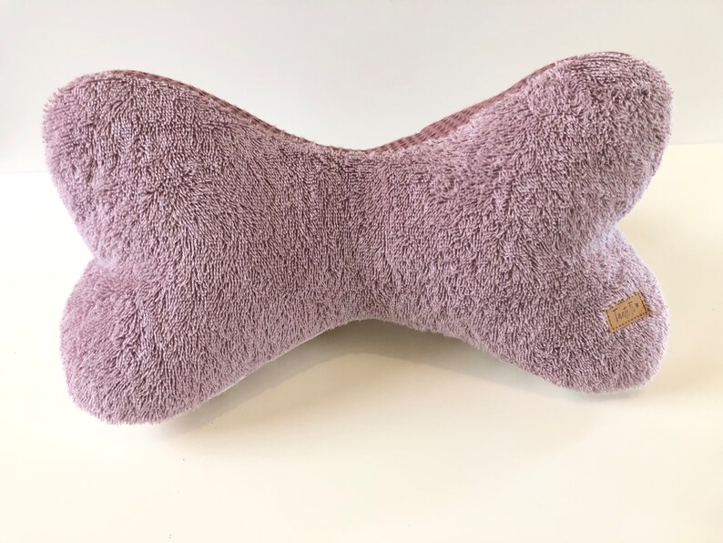 May include: A plush, mauve-colored dog bone shaped pillow. The pillow has a brown tag with the word "Made By" on it.