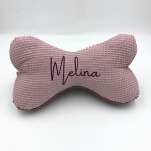 May include: A pink waffle-textured dog bone shaped pillow with the name "Melina" embroidered in purple.