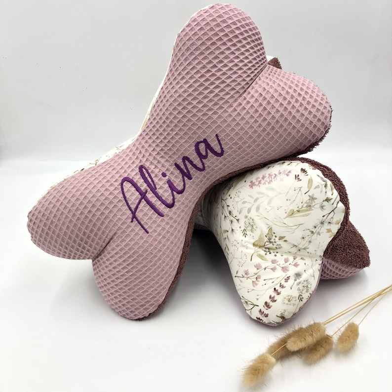 May include: Two pink and white bone-shaped pillows with a floral pattern. The top pillow has the name "Alina" embroidered on it in purple thread.