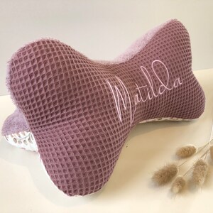 May include: A mauve-colored waffle-textured pillow shaped like a bone with the name "Matilda" embroidered in white on the side.