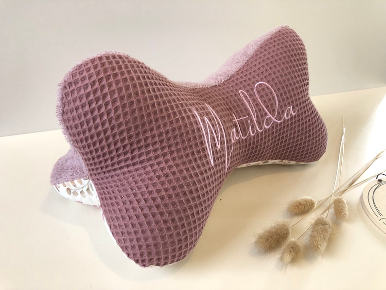 May include: A mauve-colored waffle-textured pillow shaped like a bone with the name "Matilda" embroidered in white on the side.