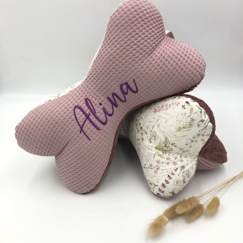 May include: Two pink and white bone-shaped pillows with a floral pattern. The top pillow has the name "Alina" embroidered on it in purple thread.