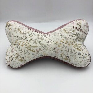May include: A pink and white floral patterned pillow with a unique bowtie shape. The pillow is made of soft fabric and has a subtle sheen.