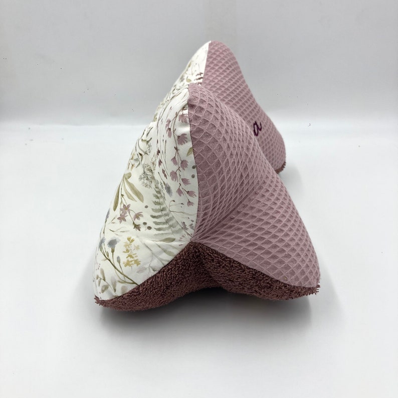 May include: A pink and white wedge-shaped pillow with a floral pattern on one side and a waffle texture on the other side. The pillow has a small embroidered "O" on the pink side.