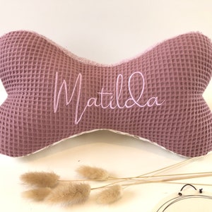 May include: A pink waffle-textured bone-shaped pillow with the name "Matilda" embroidered in white.