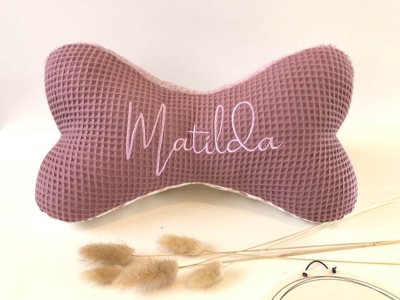 May include: A pink waffle-textured bone-shaped pillow with the name "Matilda" embroidered in white.