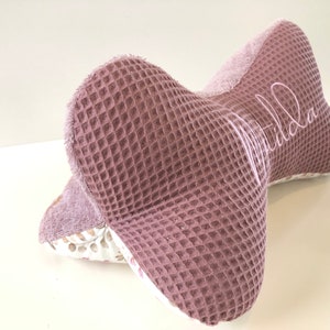 May include: A pink waffle-textured pillow with a white floral pattern on the back. The pillow is shaped like a bow and has the name "Matilda" embroidered on the front.