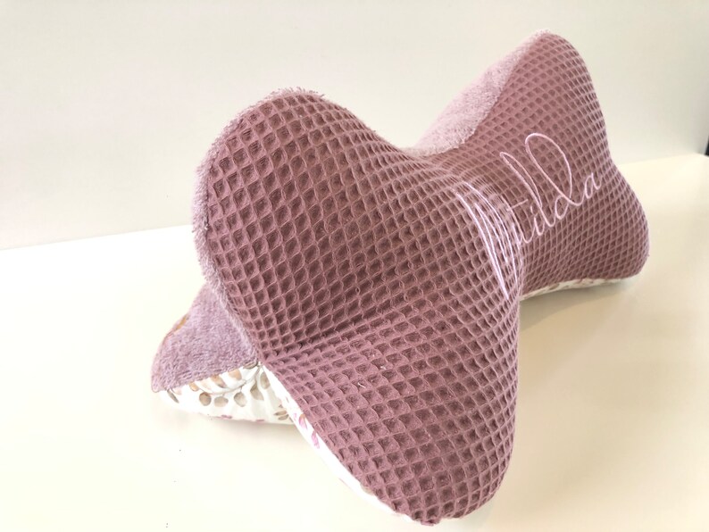 May include: A pink waffle-textured pillow with a white floral pattern on the back. The pillow is shaped like a bow and has the name "Matilda" embroidered on the front.
