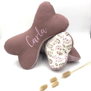 May include: A pink bone-shaped pillow with the name "Carla" embroidered on it. The pillow is made of a soft, textured fabric and is layered on top of a smaller bone-shaped pillow with a floral print.