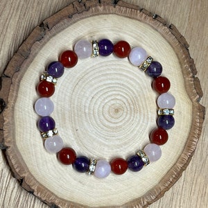 Könnte beinhalten: Ein Armband aus roten, rosa und violetten Perlen mit goldenen Akzenten. Das Armband liegt auf einer Holzoberfläche.