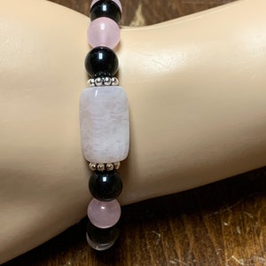 May include: A bracelet made with black and pink beads. The bracelet has a larger, rectangular pink bead in the center.