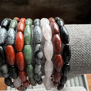 Puede incluir: Siete pulseras hechas de diferentes tipos de piedras. Las pulseras están apiladas sobre una superficie de tela gris. Las piedras son negras, rojas, verdes, grises y rosas.