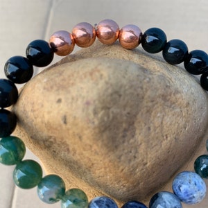 Puede incluir: Un brazalete de cuentas con piedras de &aacute;gata negra, verde y azul, y cuentas de cobre. El brazalete est&aacute; sobre una roca marr&oacute;n.