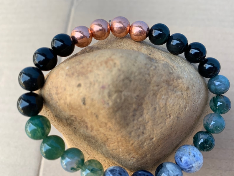Puede incluir: Un brazalete de cuentas con piedras de &aacute;gata negra, verde y azul, y cuentas de cobre. El brazalete est&aacute; sobre una roca marr&oacute;n.