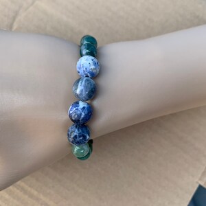 Puede incluir: Un brazalete de piedras azules y blancas con detalles de piedras verdes. El brazalete est&aacute; hecho de cuentas redondas y se lleva en la mu&ntilde;eca.