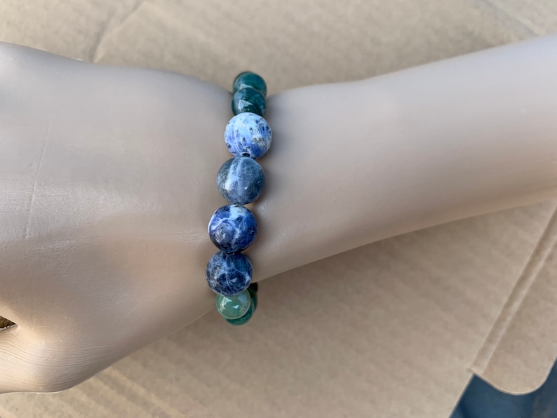 Puede incluir: Un brazalete de piedras azules y blancas con detalles de piedras verdes. El brazalete est&aacute; hecho de cuentas redondas y se lleva en la mu&ntilde;eca.