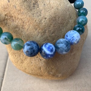 Puede incluir: Un brazalete de cuentas con piedras verdes y azules. El brazalete est&aacute; hecho de cuentas redondas con un degradado de verde a azul.