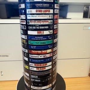 May include: A tall, cylindrical tumbler featuring a collage of album covers from various artists, including David Bowie, Cyndi Lauper, and Madonna. The design incorporates a blue rim and a black base, creating a vibrant and musical theme. The tumbler is designed to hold beverages.