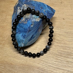 May include: Black and grey gemstone beaded bracelet. The bracelet is made with round beads and is lying on a blue and grey rock.