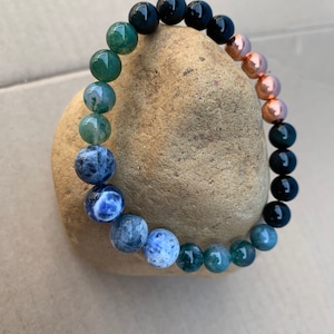 Puede incluir: Un brazalete de cuentas con cuentas negras, verdes, azules y de cobre. El brazalete est&aacute; apoyado en una roca marr&oacute;n.