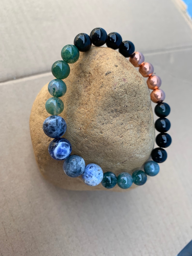 Puede incluir: Un brazalete de cuentas con cuentas negras, verdes, azules y de cobre. El brazalete est&aacute; apoyado en una roca marr&oacute;n.