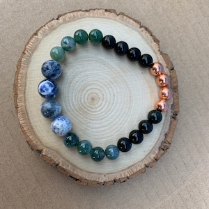 Puede incluir: Un brazalete hecho con cuentas verdes, azules, negras y de cobre. Las cuentas est&aacute;n dispuestas en un patr&oacute;n de degradado, comenzando con verde y terminando con cobre.