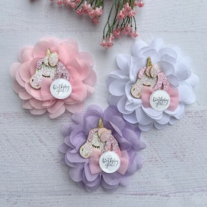 May include: Three fabric flower hair clips with a unicorn head and a pink bow. Each flower is a different color: pink, white, and purple. The center of each flower has a white circle with the text "birthday girl" in black.