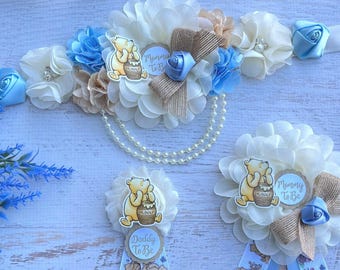 Puede incluir: Una banda de baby shower con temática de Winnie the Pooh con flores azules y blancas, un collar de perlas y un personaje de Winnie the Pooh sosteniendo un tarro de miel. La banda dice "Mommy to Be".