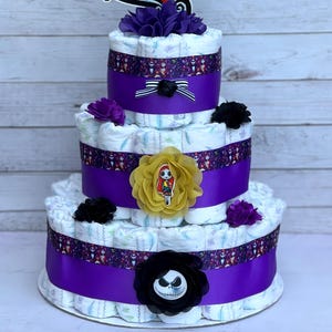 Puede incluir: Pastel de pa&ntilde;ales de tres niveles decorado con cinta morada, cinta blanca y negra y varios adornos florales. El pastel presenta un personaje de Jack Skellington y un personaje de Sally de la pel&iacute;cula Pesadilla antes de Navidad.
