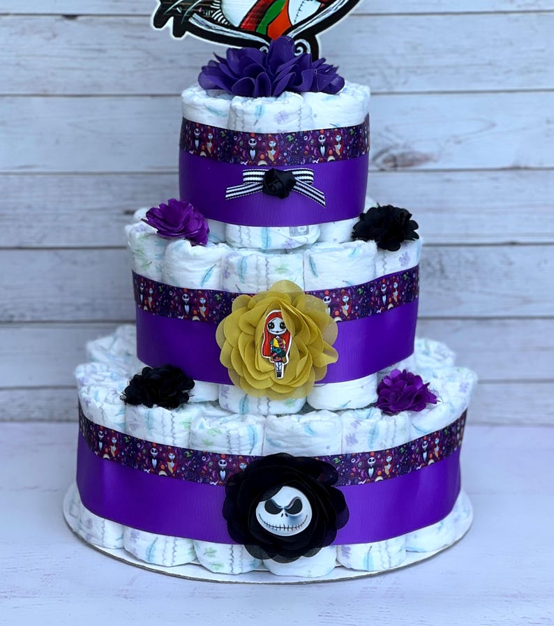 Puede incluir: Pastel de pa&ntilde;ales de tres niveles decorado con cinta morada, cinta blanca y negra y varios adornos florales. El pastel presenta un personaje de Jack Skellington y un personaje de Sally de la pel&iacute;cula Pesadilla antes de Navidad.