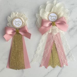 Puede incluir: Dos insignias decorativas con flores de tela color crema, lazos de satén rosa y detalles de purpurina dorada. La insignia más pequeña tiene una etiqueta que dice Daddy To Be y la más grande dice Mommy To Be.