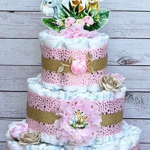 May include: A three-tiered diaper cake decorated with pink and gold ribbon, featuring a giraffe, lion, and elephant topper. The cake is adorned with pink flowers and gold glitter.