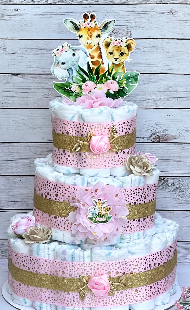 May include: A three-tiered diaper cake decorated with pink and gold ribbon, featuring a giraffe, lion, and elephant topper. The cake is adorned with pink flowers and gold glitter.