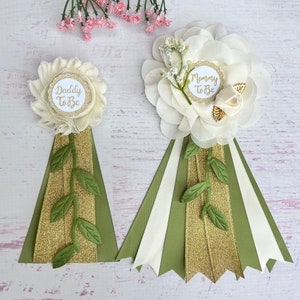 May include: Two white flower corsages with green leaves and gold glitter ribbon. One corsage says "Mommy To Be" and the other says "Daddy To Be".