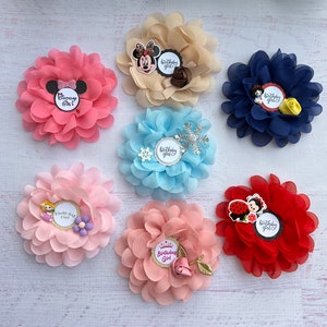 May include: A selection of fabric flower hair accessories in various colours. Each flower has a layered design and a decorative element, with tags reading "Birthday Girl" or "Birthday One". Colours include pink, blue, beige, and red.