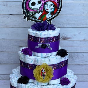Puede incluir: Un pastel de pa&ntilde;ales de tres niveles decorado con cinta morada y negra, flores y un adorno con el tema de Jack y Sally. El pastel est&aacute; hecho de pa&ntilde;ales y es un regalo popular para baby shower.