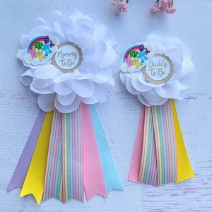 May include: Two white flower-shaped fabric pins with rainbow Care Bear designs and ribbons. One pin says "Mommy To Be" and the other says "Daddy To Be".