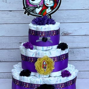 Puede incluir: Un pastel de pa&ntilde;ales de tres niveles decorado con cinta morada y negra, flores y una decoraci&oacute;n con el tema de Jack Skellington y Sally.