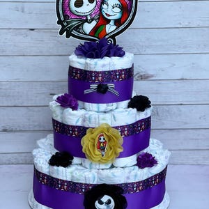 Puede incluir: Un pastel de pa&ntilde;ales de tres niveles decorado con cinta morada, cinta con estampado en blanco y negro y cubiertas de pa&ntilde;ales con estampado en blanco y negro. El pastel est&aacute; coronado con una flor morada y negra, una flor amarilla con una peque&ntilde;a figura de mu&ntilde;eca y un recorte de cart&oacute;n de Jack Skellington y Sally de Pesadilla antes de Navidad.