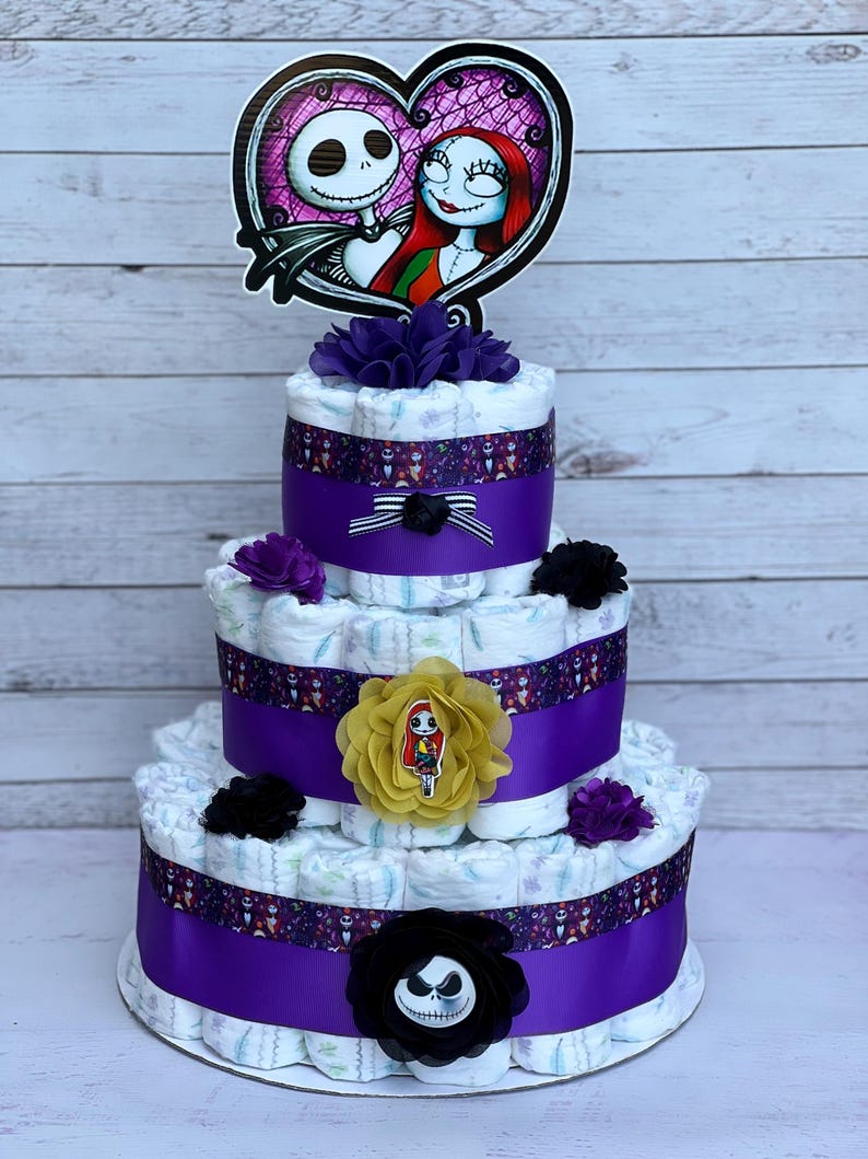 Puede incluir: Un pastel de pa&ntilde;ales de tres niveles decorado con cinta morada, cinta con estampado en blanco y negro y cubiertas de pa&ntilde;ales con estampado en blanco y negro. El pastel est&aacute; coronado con una flor morada y negra, una flor amarilla con una peque&ntilde;a figura de mu&ntilde;eca y un recorte de cart&oacute;n de Jack Skellington y Sally de Pesadilla antes de Navidad.