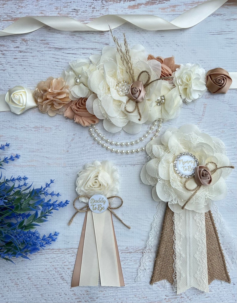 Może przedstawiać: Zestaw dw&oacute;ch kwiatowych szarf na baby shower. Szarfy wykonane są z tkaniny w kolorze kości słoniowej i beżowym z dodatkami z koronki i juty. Jedna szarfa ma napis "Mommy to Be", a druga "Daddy to Be".