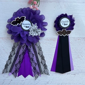 May include: Two purple and black Halloween themed baby shower boutonnieres. One says "Mommy to Be" with a bat and spider web. The other says "Daddy to Be" with a bat.